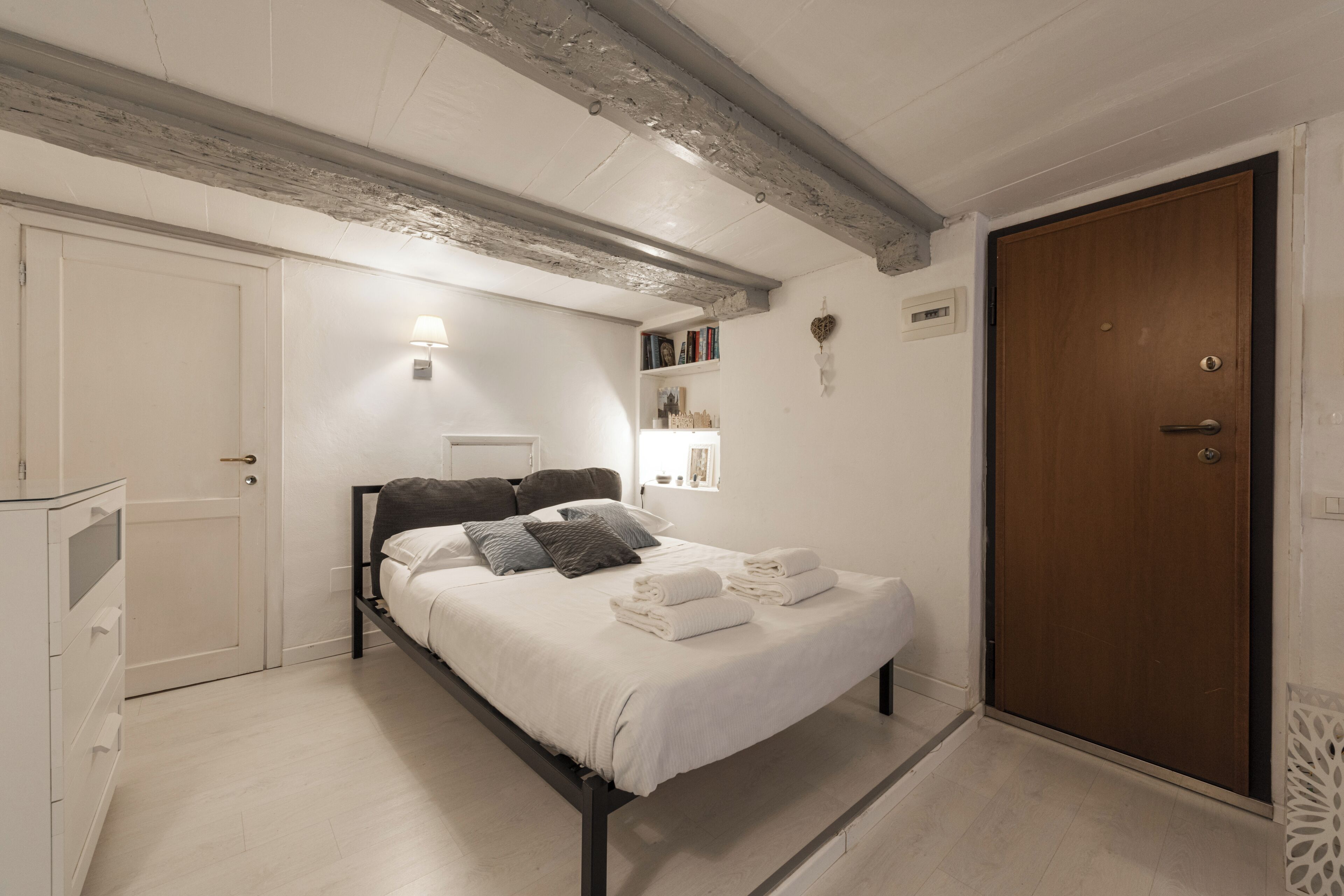 A bedroom with a bed, a wardrobe, a bedside table, a lamp, and a shelf with books.