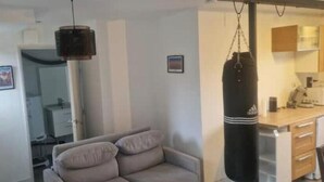 A living room with a sofa, a punching bag, a microwave, and a small kitchen area. - T2 Fully Equipped 4 Sleeps - Comfort and Proximity to Amenities (Réalmont)