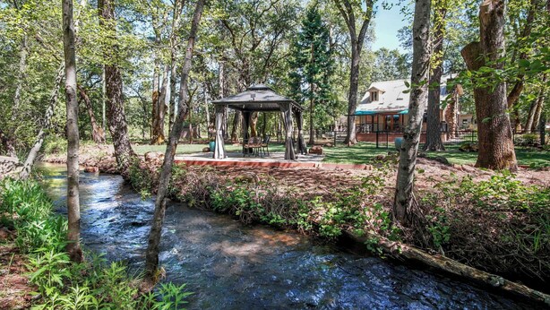 A gazebo in a forested area with a stream, surrounded by trees and a building in the background.