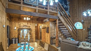 A wooden interior with a dining table, chairs, a sofa, a staircase, and a chandelier.