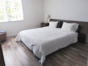Een slaapkamer met een groot bed, een nachtkastje, een lamp en een raam met jaloezieën.