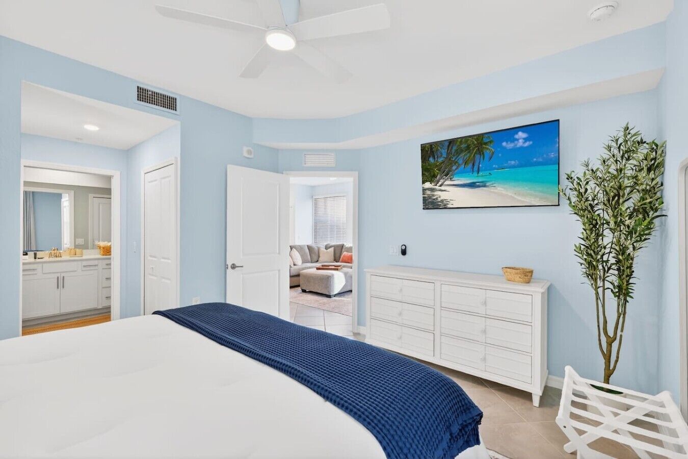 Une chambre à coucher avec un lit, une commode, un tableau représentant une plage et une plante.