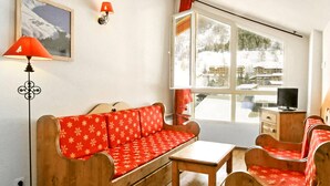 Une pièce chaleureuse avec un canapé en bois, une petite table et une chaise, le tout recouvert d’un tissu à motifs rouges. Une lampe et un téléviseur sont posés sur une commode en bois.