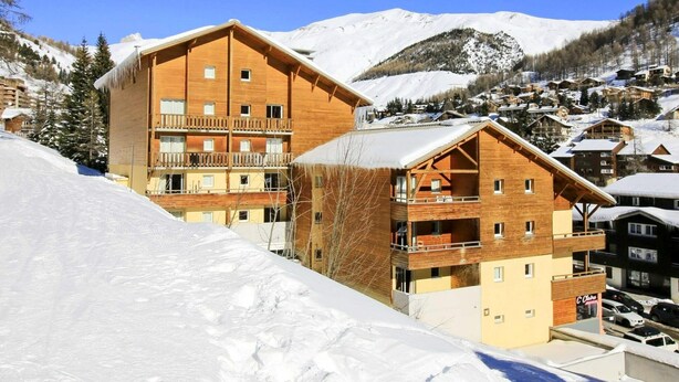 Un immeuble en bois à plusieurs étages, doté de balcons, entouré de montagnes enneigées et situé à proximité d’une station de ski.