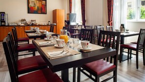 Ein Hotel-Speisesaal mit einem langen Tisch, der für ein Frühstück gedeckt ist, Holzsesseln, einer Kaffeemaschine und einem Gemälde an der Wand.