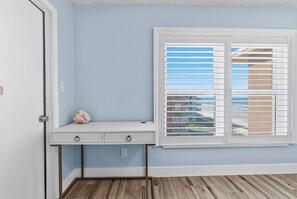 Ein Zimmer mit einem weißen Schreibtisch, einem Fenster mit Jalousien und Blick auf Gebäude.