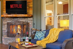 En hyggelig stue med en stenvægspejs, et fladskærms-tv der viser 'NETFLIX', og en behagelig blå sofa med et gult tæppe.