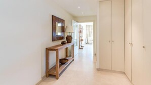 Un pasillo con una consola de madera, un espejo y una lámpara. Hay una puerta a la derecha y se ve una vista hacia otra habitación.