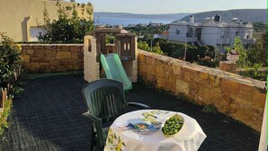 A patio with a table set for two, chairs, a small slide, and a view of the sea and buildings. - Άνετος οικογενειακός  χώρος στην Αγία Ερμιόνη Χίου λίγα λεπτά από την παραλία! (AGIA ERMIONI)