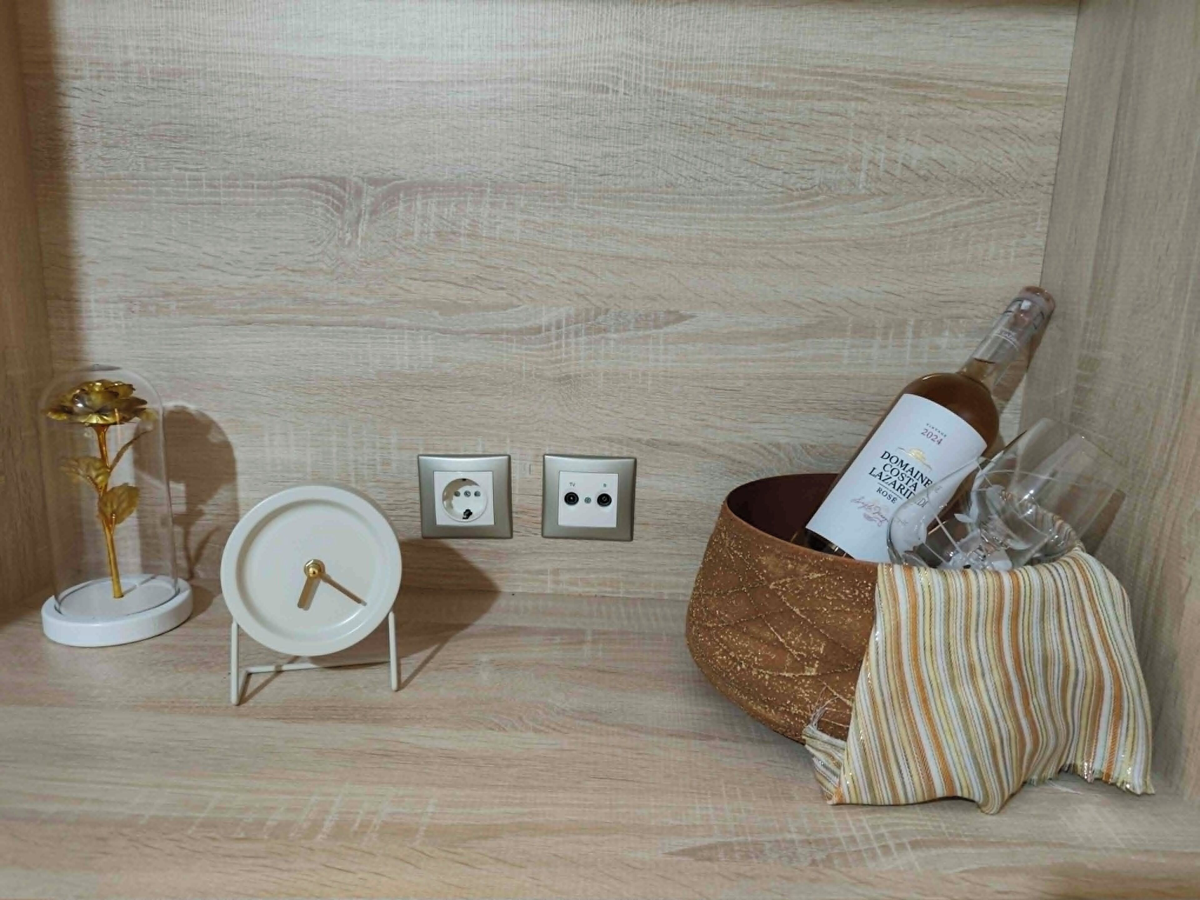 Pareti in legno con due prese elettriche, una piccola sveglia bianca, un fiore decorativo e una bottiglia di vino in un cesto di vimini.