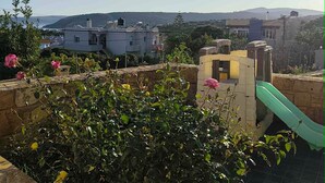 A residential area with a green slide, stone walls, and a view of distant buildings and hills. - Άνετος οικογενειακός χώρος στην Αγία Ερμιόνη Χίου λίγα λεπτά από την παραλία! (AGIA ERMIONI)