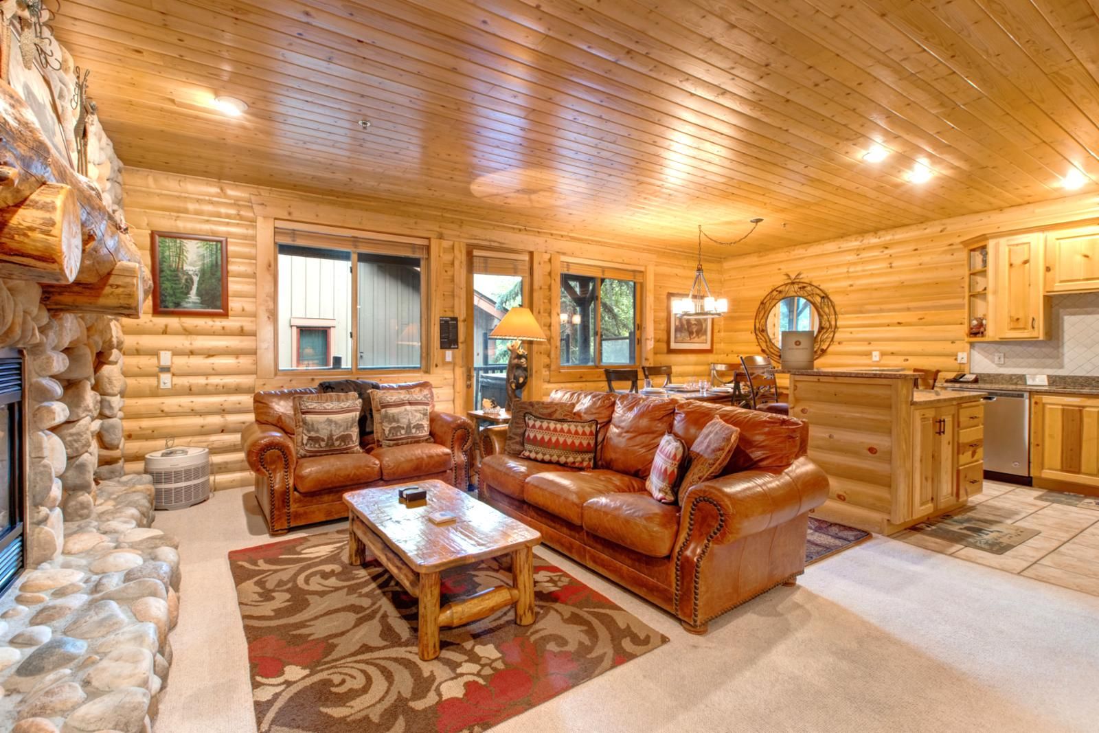 A log cabin interior with a stone fireplace, leather sofas, a dining area, and a kitchen.