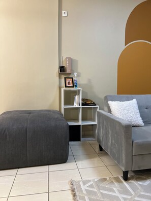 Un salon avec un canapé gris, un pouf gris, une étagère blanche et un tapis à motifs posé sur un sol carrelé.