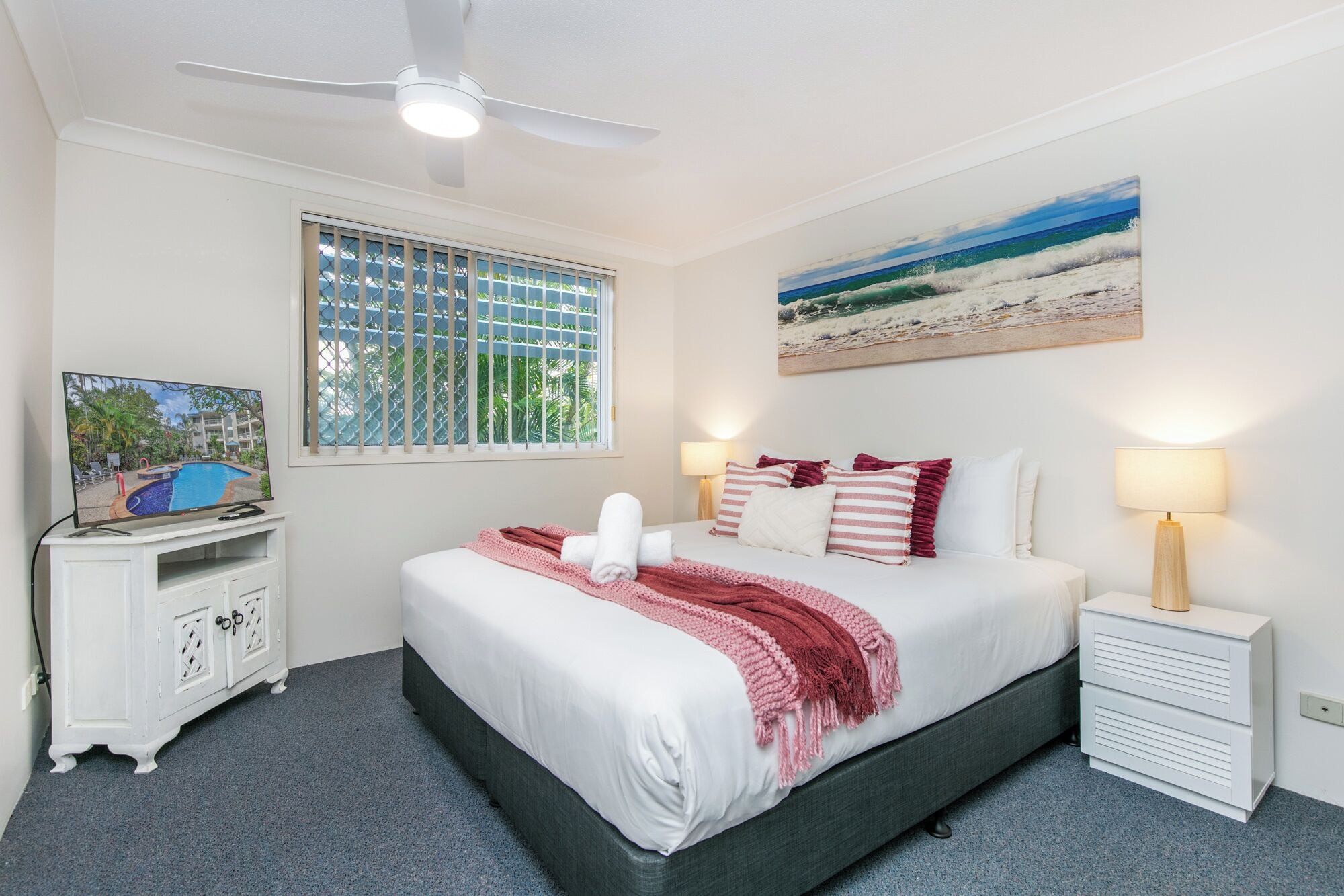 Ein Schlafzimmer mit einem Bett, einem Fernseher, einem Deckenventilator, einem Fenster mit Jalousien, einem Gemälde und einem Nachttisch.
