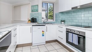 A modern kitchen with white cabinetry, a built-in dishwasher, a stove, and a window with a view of greenery. - Bliss On Beach Parade (Surfers Paradise)