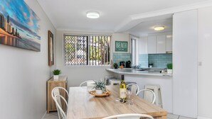 A modern kitchen with white cabinets, a wooden dining table, and a view of a cityscape through a window. - Bliss On Beach Parade (Surfers Paradise)