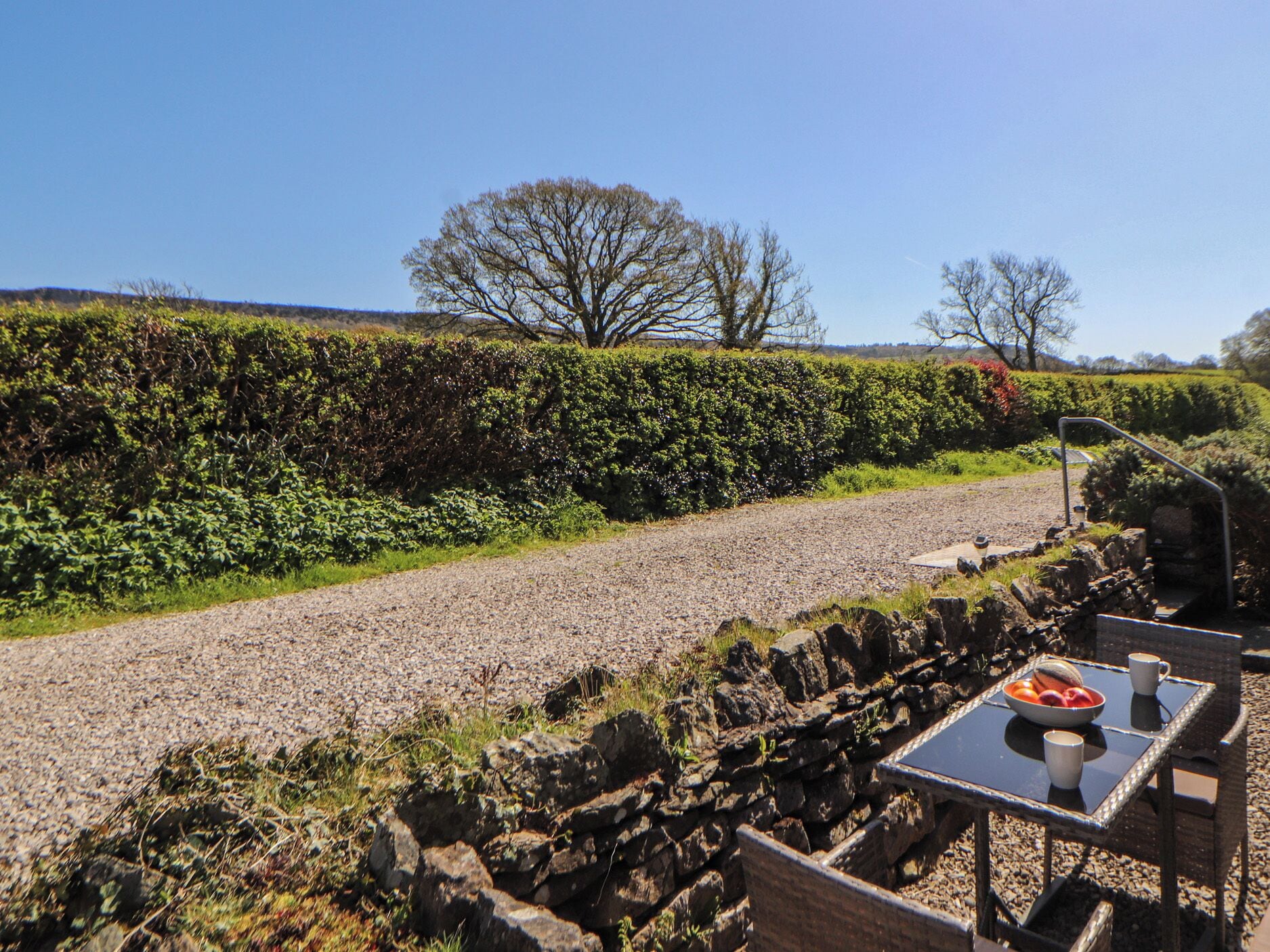 A gravel path with a stone wall and a table with a bowl of fruit and a cup.