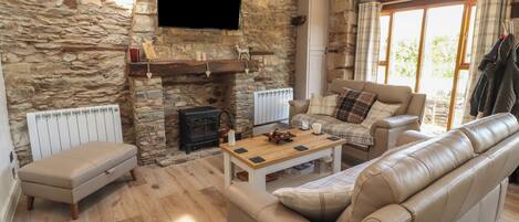 A stone-walled living room with a fireplace, leather sofas, a wooden coffee table, and a television mounted on the wall.