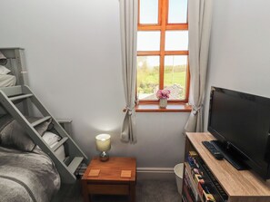 Ein Schlafzimmer mit Bett, Fernseher, einem kleinen Tisch mit Lampe, einem Fenster mit Vorhängen und einem Bücherregal.