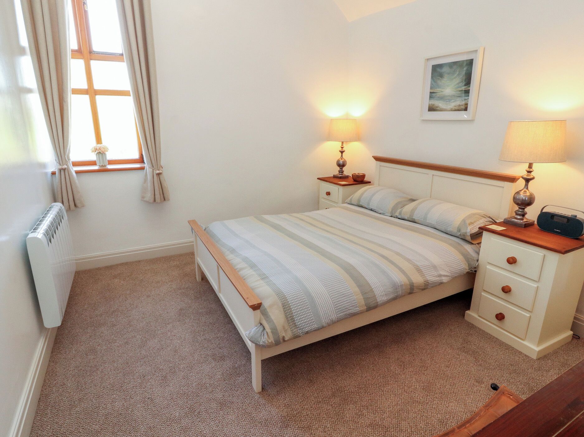 Un dormitorio con cama, mesitas de noche, lámparas, un radiador, una ventana con cortinas y un cuadro en la pared.