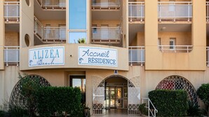 Um edifício com vários andares, varandas, uma placa com a inscrição 'Alizea Beach Residence' e uma entrada com portão.