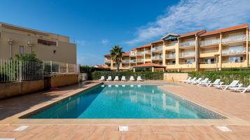 A swimming pool with lounge chairs, a building with balconies, and a clear sky.