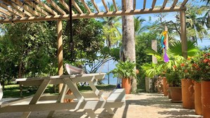 A wooden pergola with a picnic table, surrounded by potted plants and palm trees.