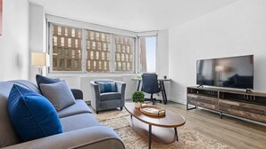 A modern living room with a sofa, armchair, coffee table, TV, and a large window with blinds. - Spacious Apt in Murray Hill (New York, NY)