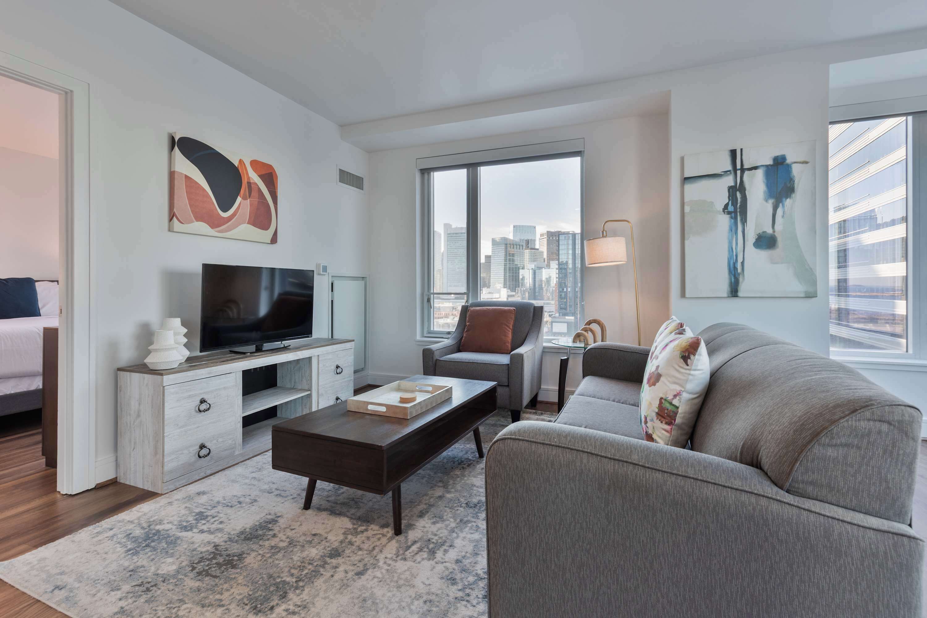 A modern living room with a flat-screen TV, grey sofa, and a city view through large windows.