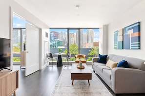 Ein modernes Wohnzimmer mit einer Couch, einem Couchtisch und Sesseln, großen Fenstern mit Blick auf die Stadt und Kunstwerken an der Wand.