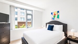A modern hotel room with a large bed, a dresser, a TV, and a window with a view of buildings. - Luxury Apt On Canal in Cambridge (Cambridge, MA)