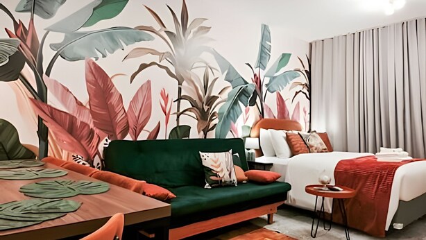 A hotel room with a bed, a sofa, a dining table, and a mural of tropical leaves. - Apartamento Cheio de Estilo perto da FEI (São Bernardo do Campo)