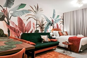 A hotel room with a bed, a sofa, a dining table, and a mural of tropical leaves. - Apartamento Cheio de Estilo perto da FEI (São Bernardo do Campo)