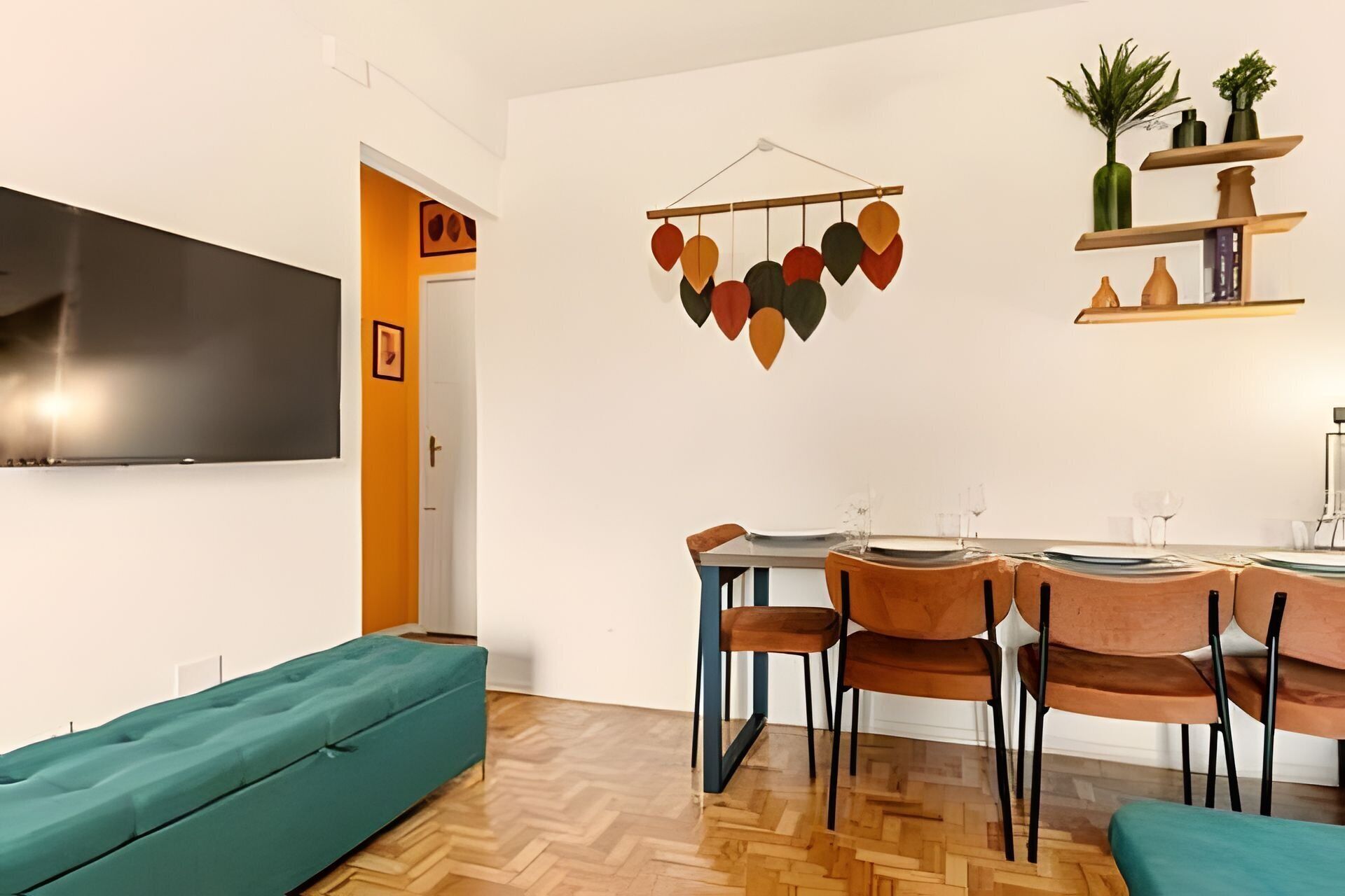 A modern living room with a sofa, a dining area with chairs and a table, a flat-screen TV mounted on the wall, and a decorative hanging piece.