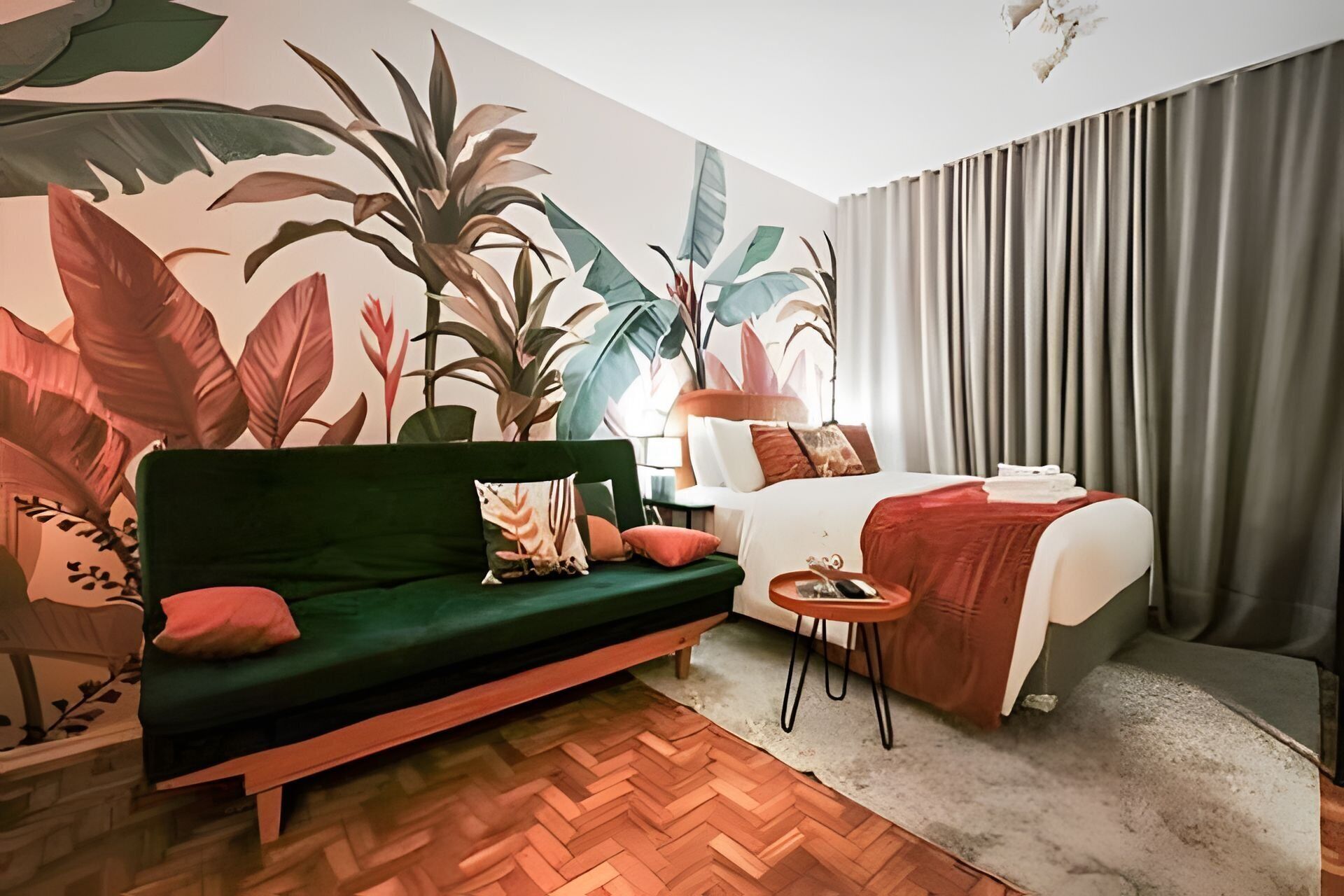 A hotel room with a green sofa, a bed with a red blanket, a small round table, a lamp, and a large wall mural of tropical leaves.