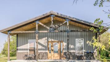 A rustic building with a wooden door, a porch with chairs, and a grill outside.