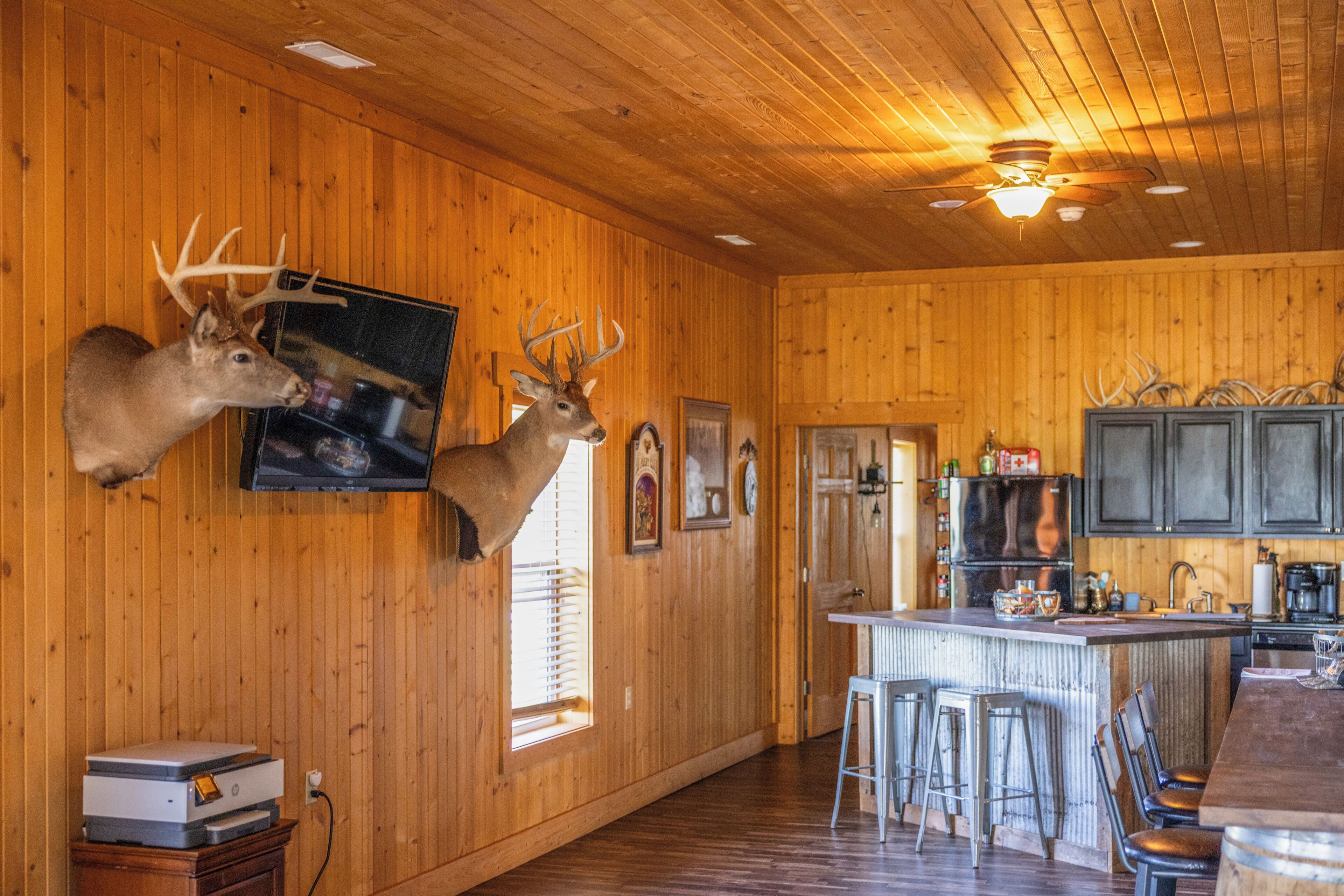 A cabin interior with wooden walls, mounted deer heads, a bar area, and a kitchen with modern appliances.