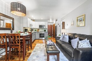 Eine moderne Küche mit weißen Schränken, ein Essbereich mit Holzsesseln, ein schwarzes Ledersofa und ein Teppich mit Muster.