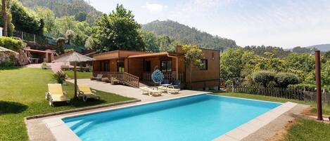 A rectangular swimming pool with lounge chairs and a wooden deck, surrounded by a grassy area and a mountain backdrop.
