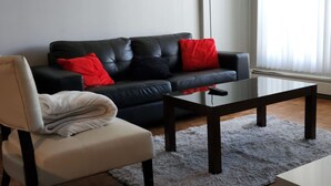Ein Wohnzimmer mit einer schwarzen Ledersofa, roten Kissen, einem Glastisch und einem weißen Sessel, sowie einem flauschigen Teppich.