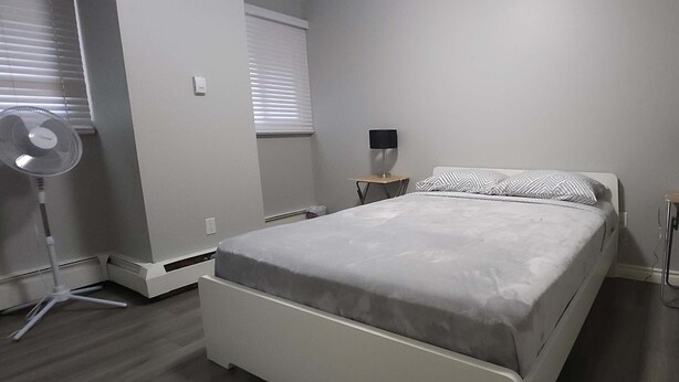 Ein Schlafzimmer mit einem Bett, Nachttischen, einem Standventilator und einem Fenster mit Jalousien.