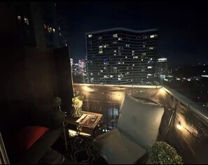 Ein Balkon mit Blick auf eine beleuchtete Stadtlandschaft bei Nacht, mit einem modernen Gebäude und erleuchteten Fenstern.