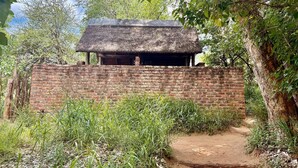Una estructura rústica con techo de paja y pared de ladrillo, rodeada de vegetación y un sendero de tierra.