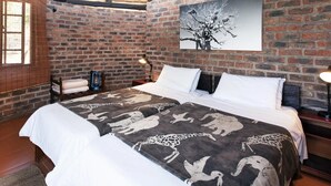 Una habitación con pared de ladrillo, cama con cubrecama estampado, un cuadro de un árbol y techo de paja.