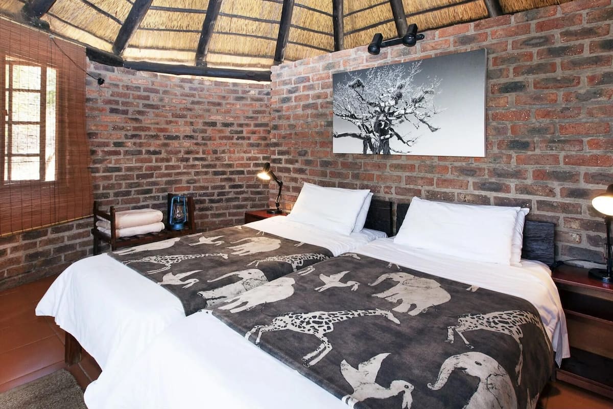 A room with a brick wall, a bed with a patterned bedspread, a painting of a tree, and a thatched roof.