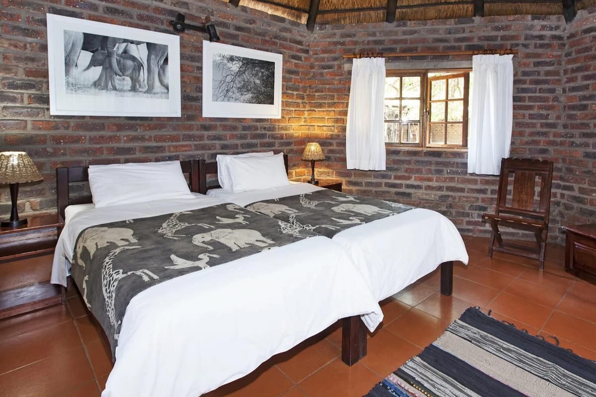 A room with a brick wall, a bed with a patterned bedspread, a window with curtains, and a wooden chair.