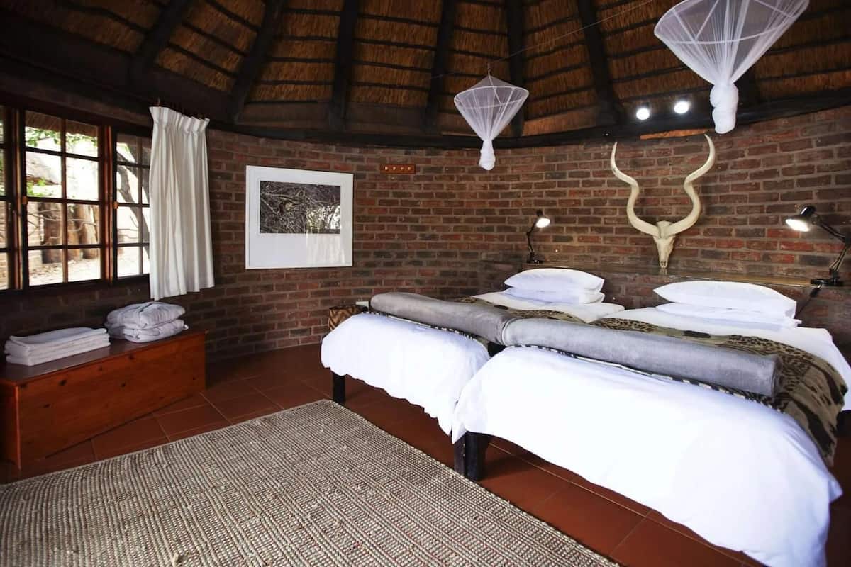 A room with a brick wall, a thatched roof, a bed with white linens, a wooden bench with towels, a framed picture, and a wall-mounted light fixture.
