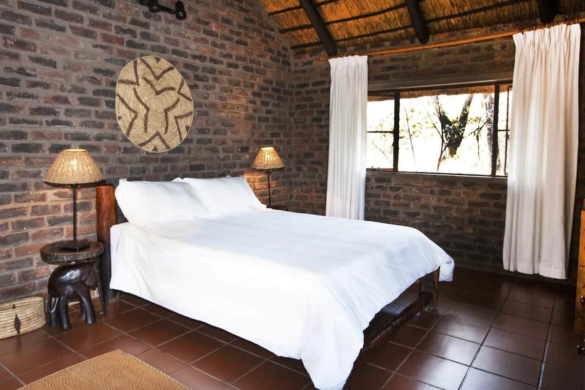 A room with a brick wall, a bed with white bedding, two lamps, and a window with curtains.