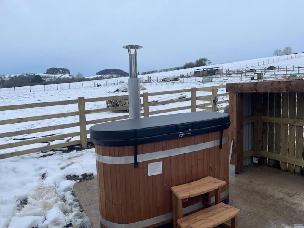 A wooden sauna with a bench, a metal pole, and a wooden fence in a snowy landscape.