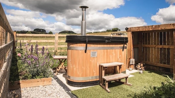 A wooden hot tub with a metal chimney, surrounded by a wooden deck and a garden with purple flowers.